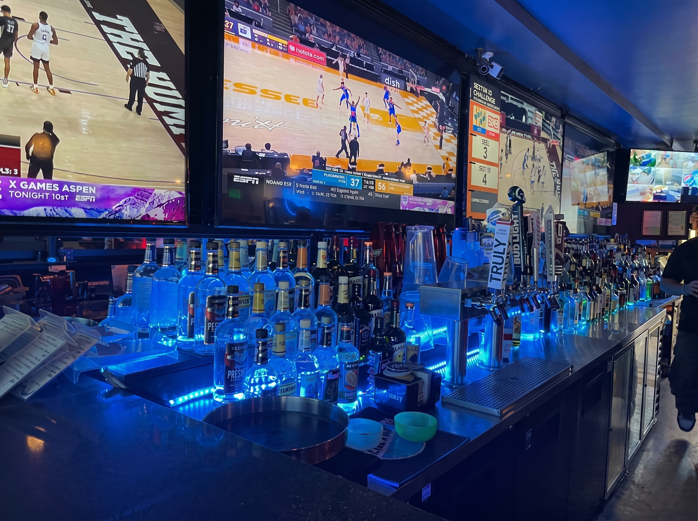 The back bar at The Blue Moon Lounge — full liquor lineup under blue light, games on every screen